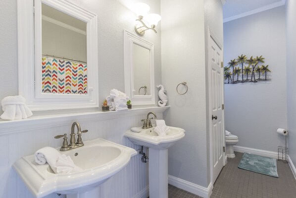 A bathroom with two sinks, a mirror, and a toilet. There is a towel rack and a small shelf with toiletries.