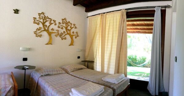 A room with two beds, a small table, a wall-mounted lamp, and a window with curtains.