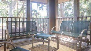 A covered outdoor seating area with two green cushioned chairs and a bench, surrounded by wooden railings and large windows overlooking trees.