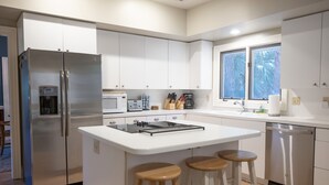 A modern kitchen with white cabinetry, a stainless steel refrigerator, a built-in microwave, a coffee maker, and a dishwasher. There are two wooden stools at the island.