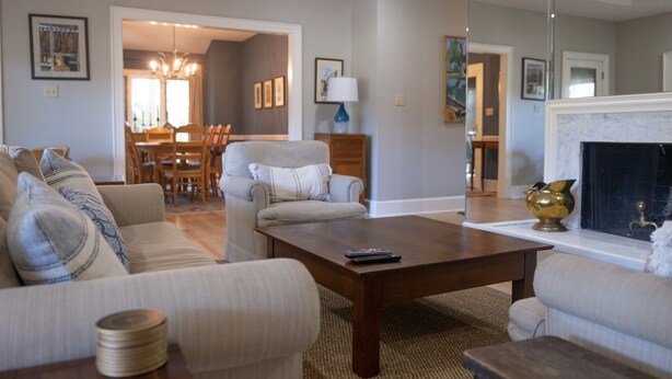 A living room with a sofa, coffee table, and fireplace. There is a dining area in the background with a chandelier.