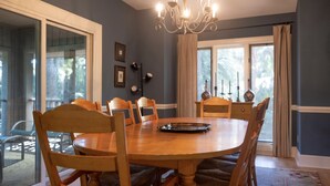 A dining room with a round wooden table, six chairs, a chandelier, and a view through large windows.