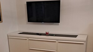 A room with a white cabinet, a flat-screen TV mounted on the wall, and a round table with a geometric-patterned tablecloth.