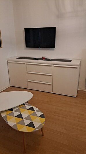 A room with a white cabinet, a flat-screen TV mounted on the wall, and a round table with a geometric-patterned tablecloth.