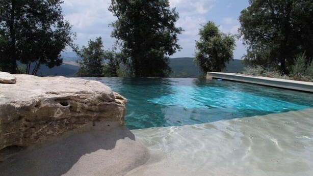 Piscina cristalina com borda rochosa e vista para árvores e colinas ao fundo.