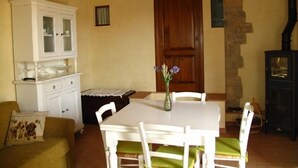 Uma área de refeições com mesa e cadeiras brancas, um armário com portas de vidro, uma porta de madeira e uma parede de pedra.