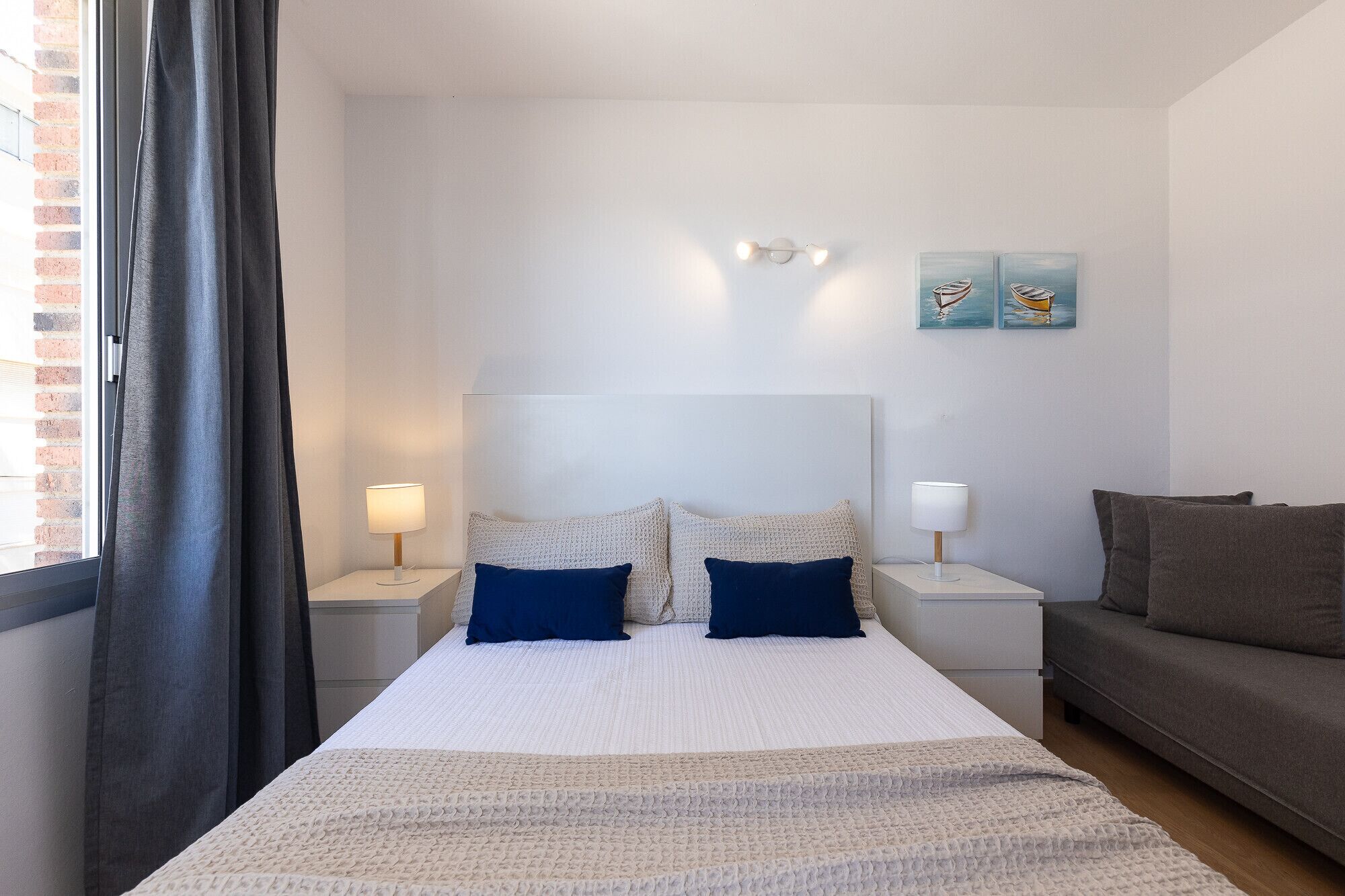 A bedroom with a large bed, two bedside tables with lamps, a grey sofa, and two wall-mounted paintings.