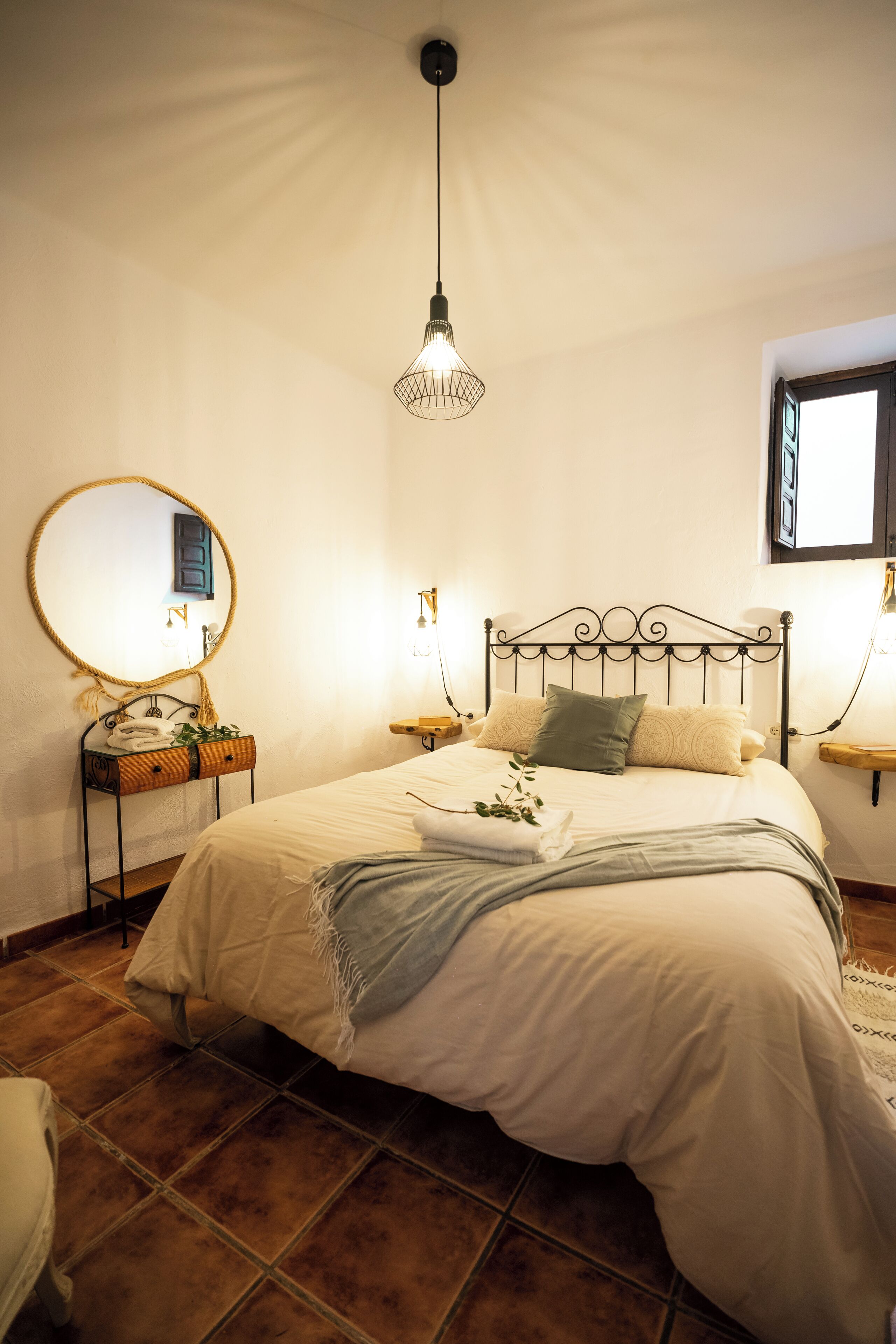 Un lit bien fait, avec une tête de lit, une table de chevet dotée d’un miroir et un luminaire suspendu.