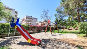 Ein Spielplatz mit Rutsche und Schaukeln, umgeben von Grünflächen und Gebäuden.