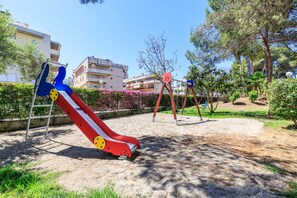A playground with a slide and swings, surrounded by greenery and buildings.