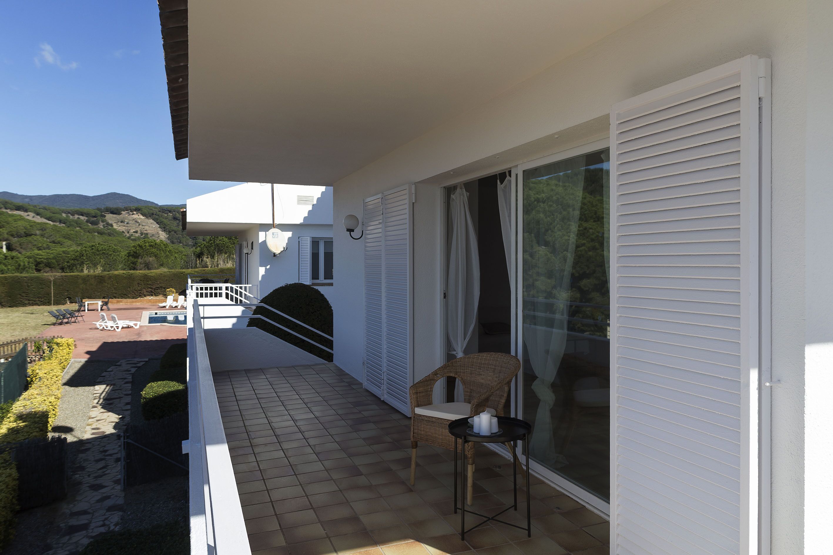 A balcony with a wicker chair and a small table, overlooking a pool and a garden.