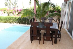A wooden table and chairs set up by a poolside, with a palm tree and flowering plants in the background.