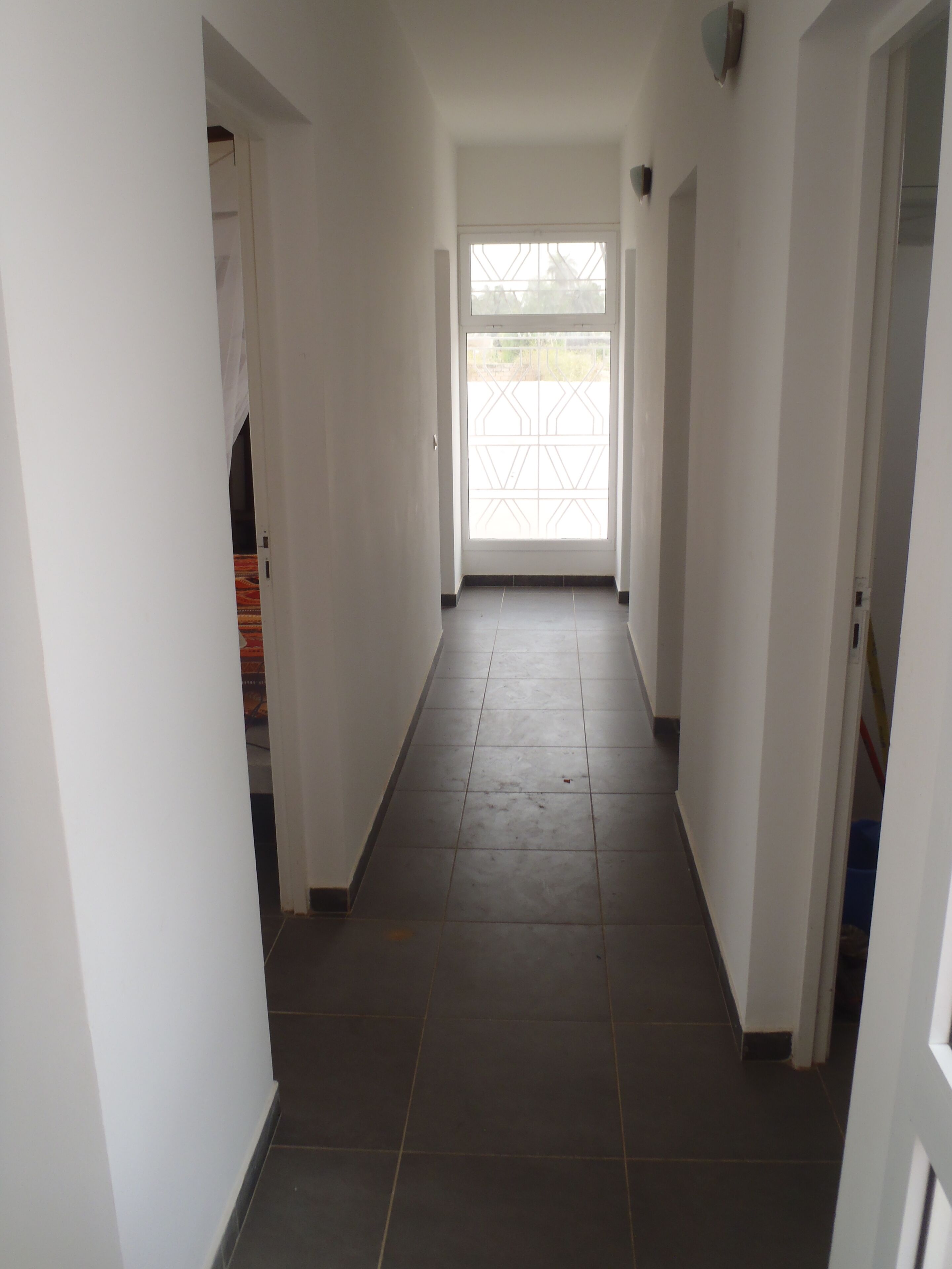 A narrow hallway with white walls and a tiled floor, leading to a large glass door at the end.