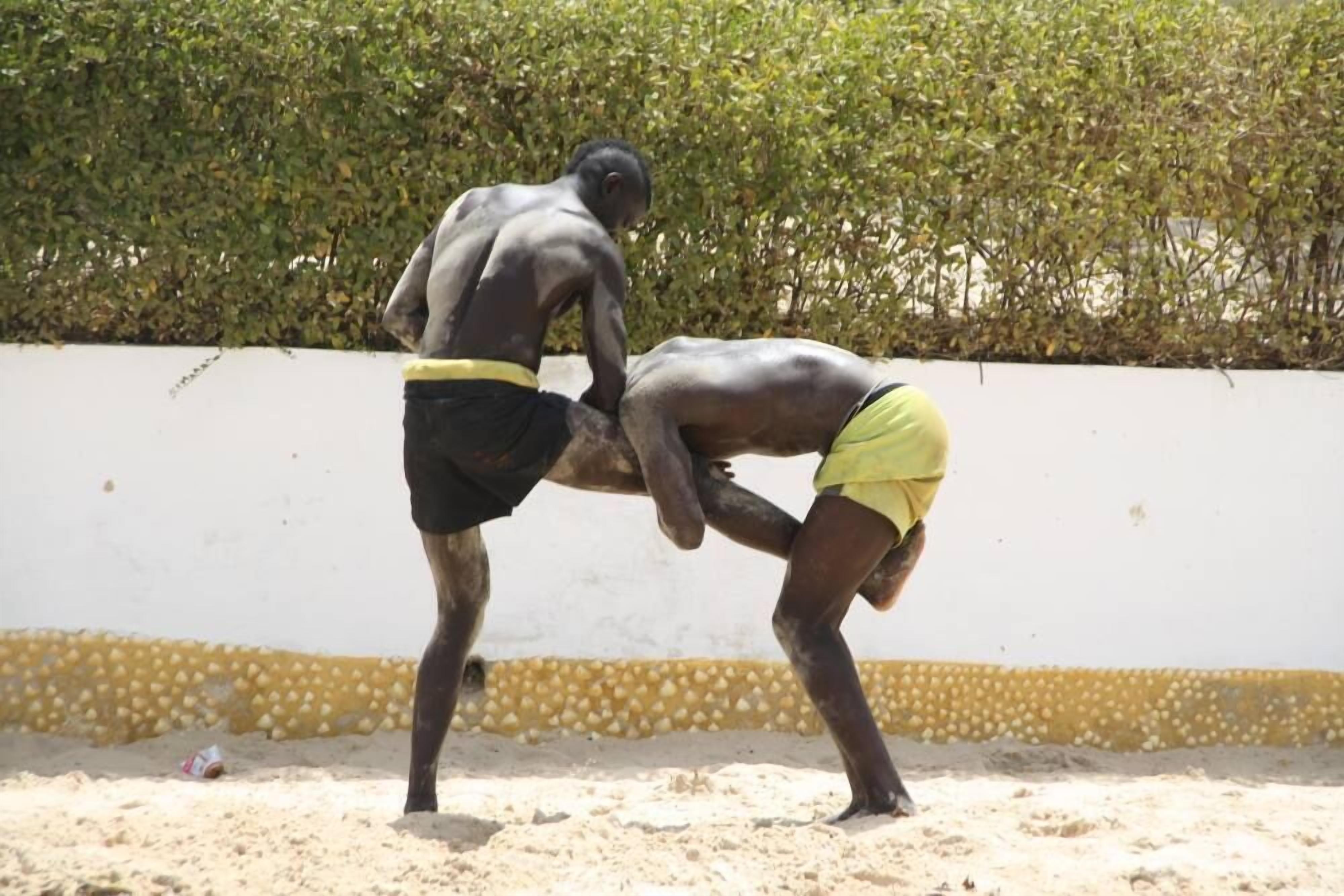 Two individuals engaged in a physical activity on a sandy surface with a white wall and green foliage in the background.