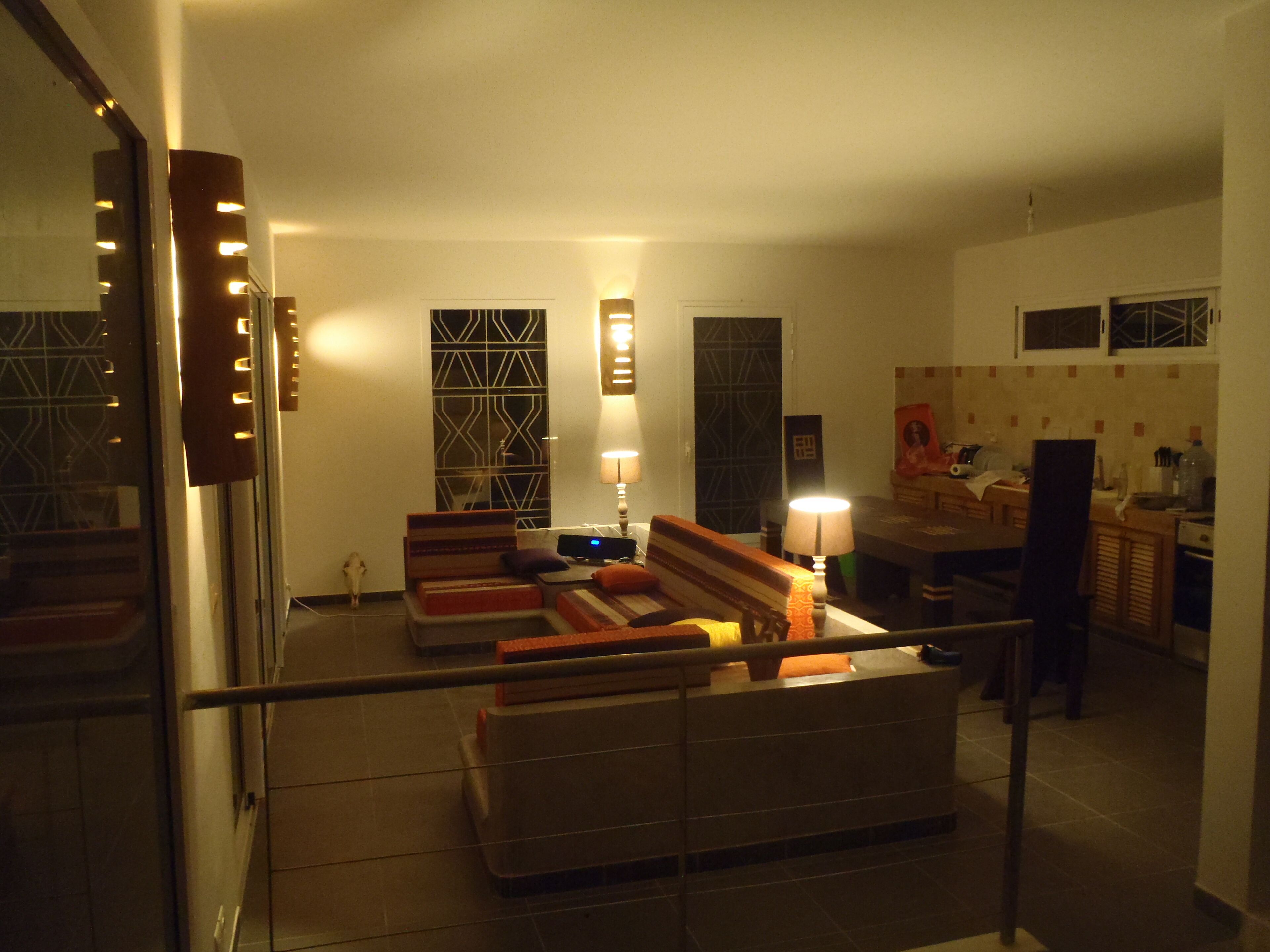 A living room with a sofa set, a coffee table, and a lamp. There is a window with a decorative screen, a door with a mirror, and a kitchen area in the background.