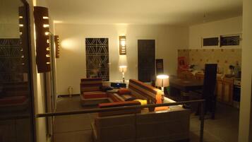A living room with a sofa set, a coffee table, and a lamp. There is a window with a decorative screen, a door with a mirror, and a kitchen area in the background.