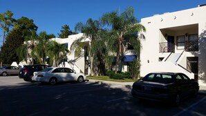 Un estacionamiento con varios autos, un edificio blanco con balcón y palmeras.