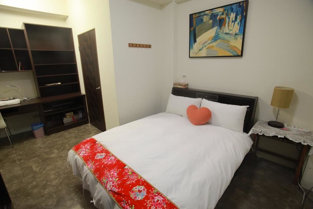 A bedroom with a bed, a desk, and a chair. There is a painting on the wall and a lamp on a side table.