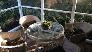 A glass-top table surrounded by wicker chairs in a sunlit room with a view of greenery outside.