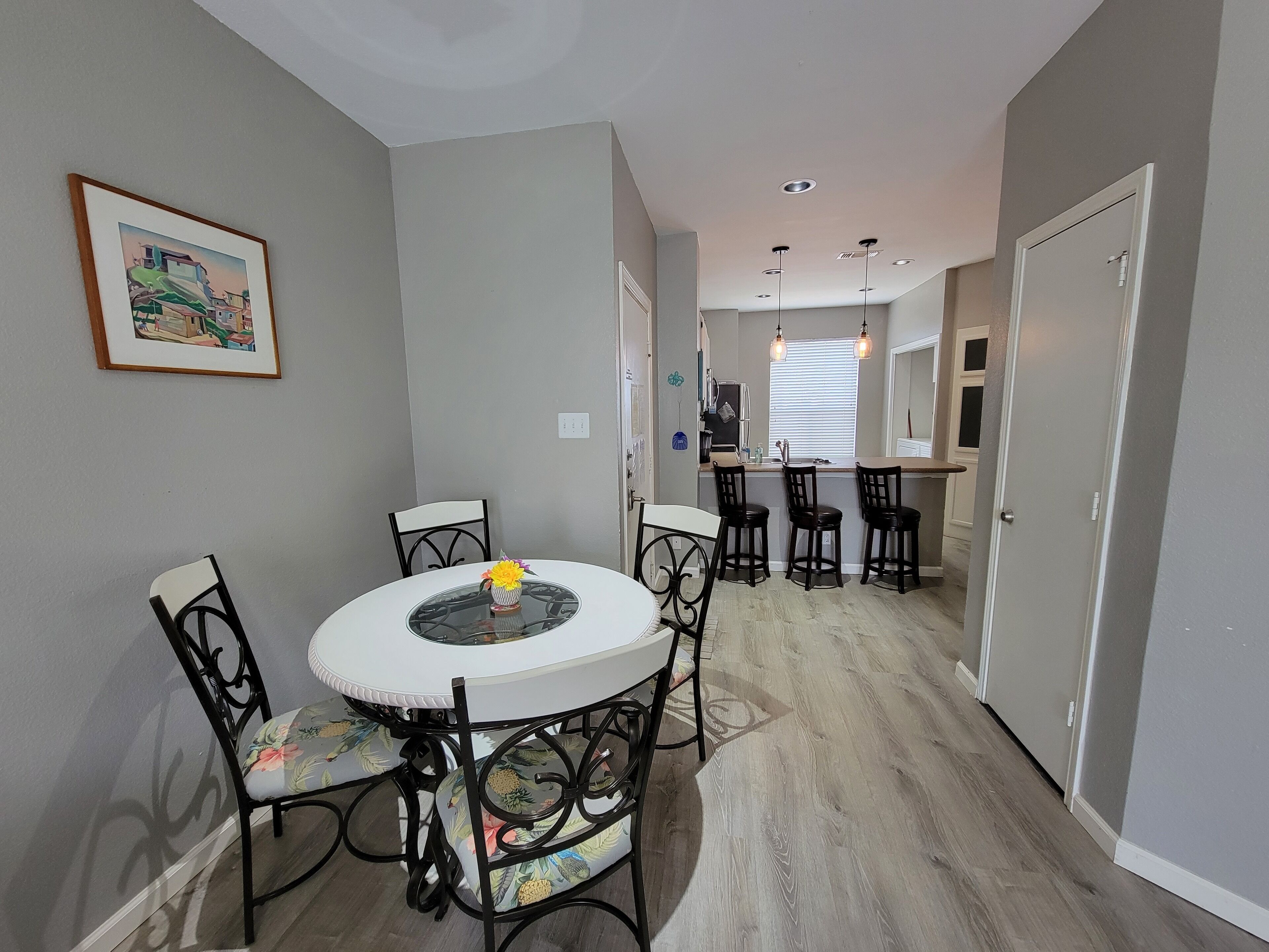 A dining area with a round table and four chairs, a kitchen in the background, and a hallway leading to a door.