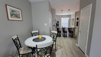 A dining area with a round table and four chairs, a kitchen in the background, and a hallway leading to a door.