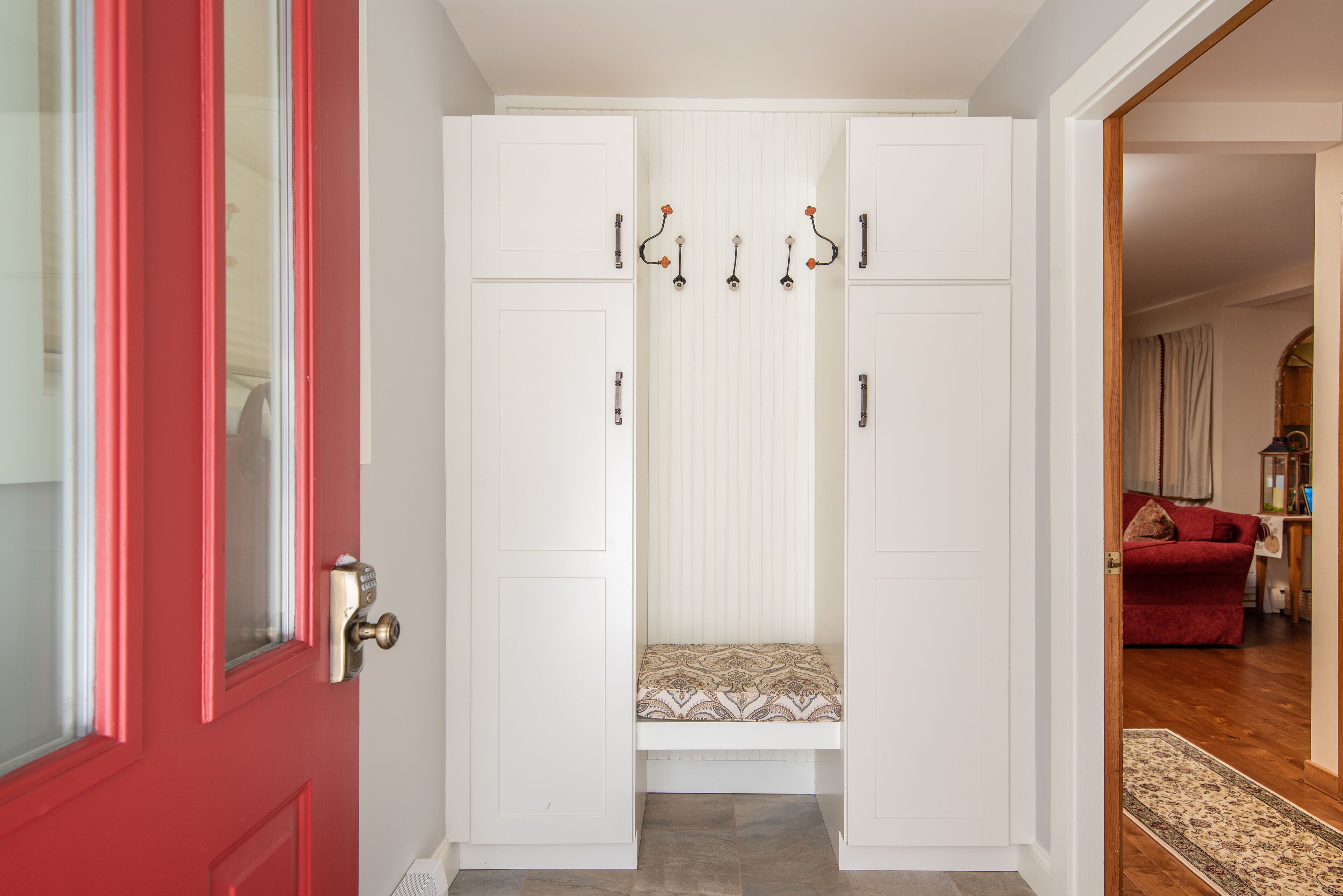 Un couloir avec une porte rouge, un banc blanc orné de coussins à motifs et un plancher en bois.