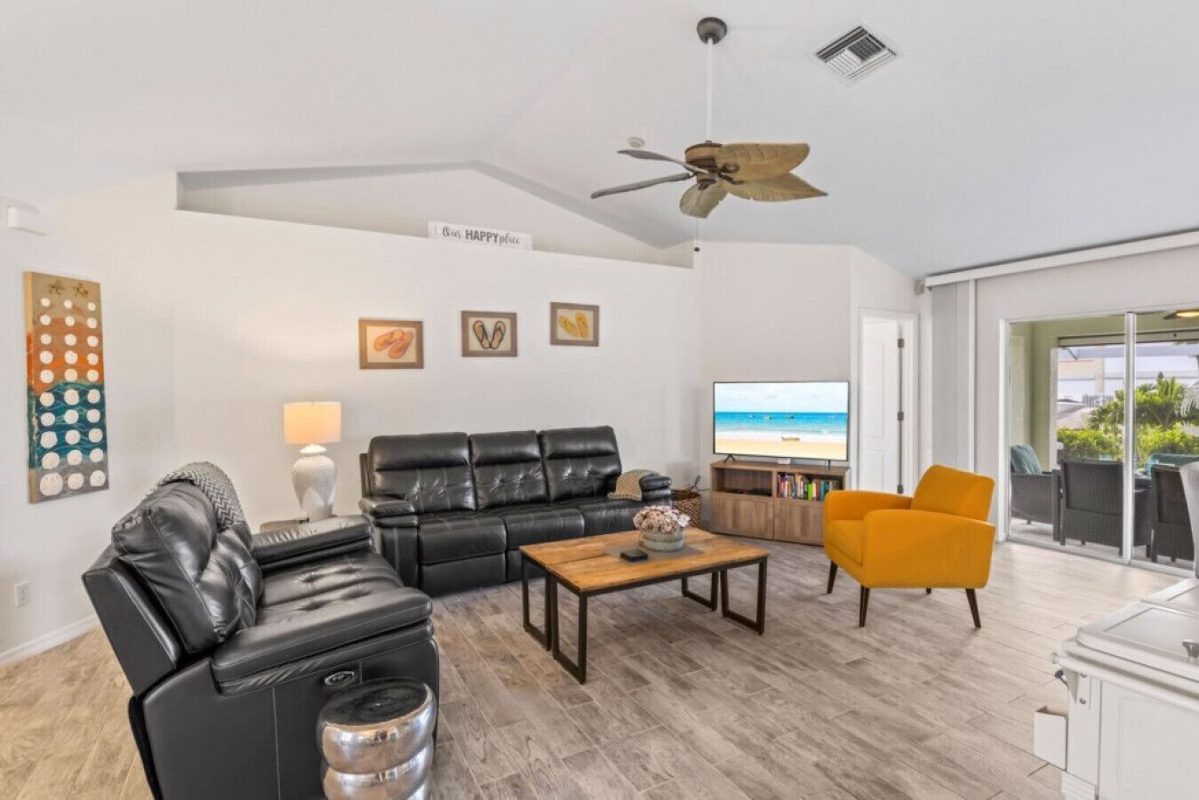 Una sala de estar moderna con un sofá de cuero negro, una mesa de centro de madera, un sillón amarillo y un televisor que muestra una escena de playa.