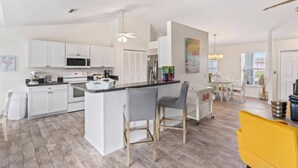 Una cocina moderna con armarios blancos, una isla central y un comedor con una silla amarilla.