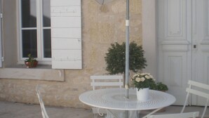 A white metal table and chairs set under a white umbrella, with a potted plant and a small window with shutters.
