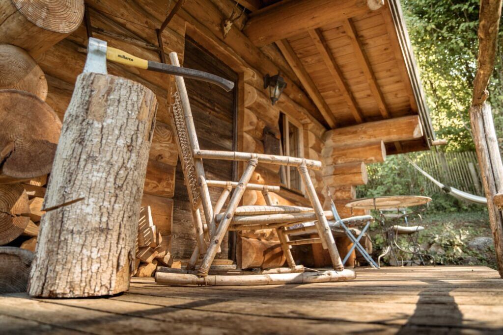 A rustic wooden cabin with a large log pile, a metal axe, and a wooden chair outside.