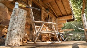 A rustic wooden cabin with a large log pile, a metal axe, and a wooden chair outside.