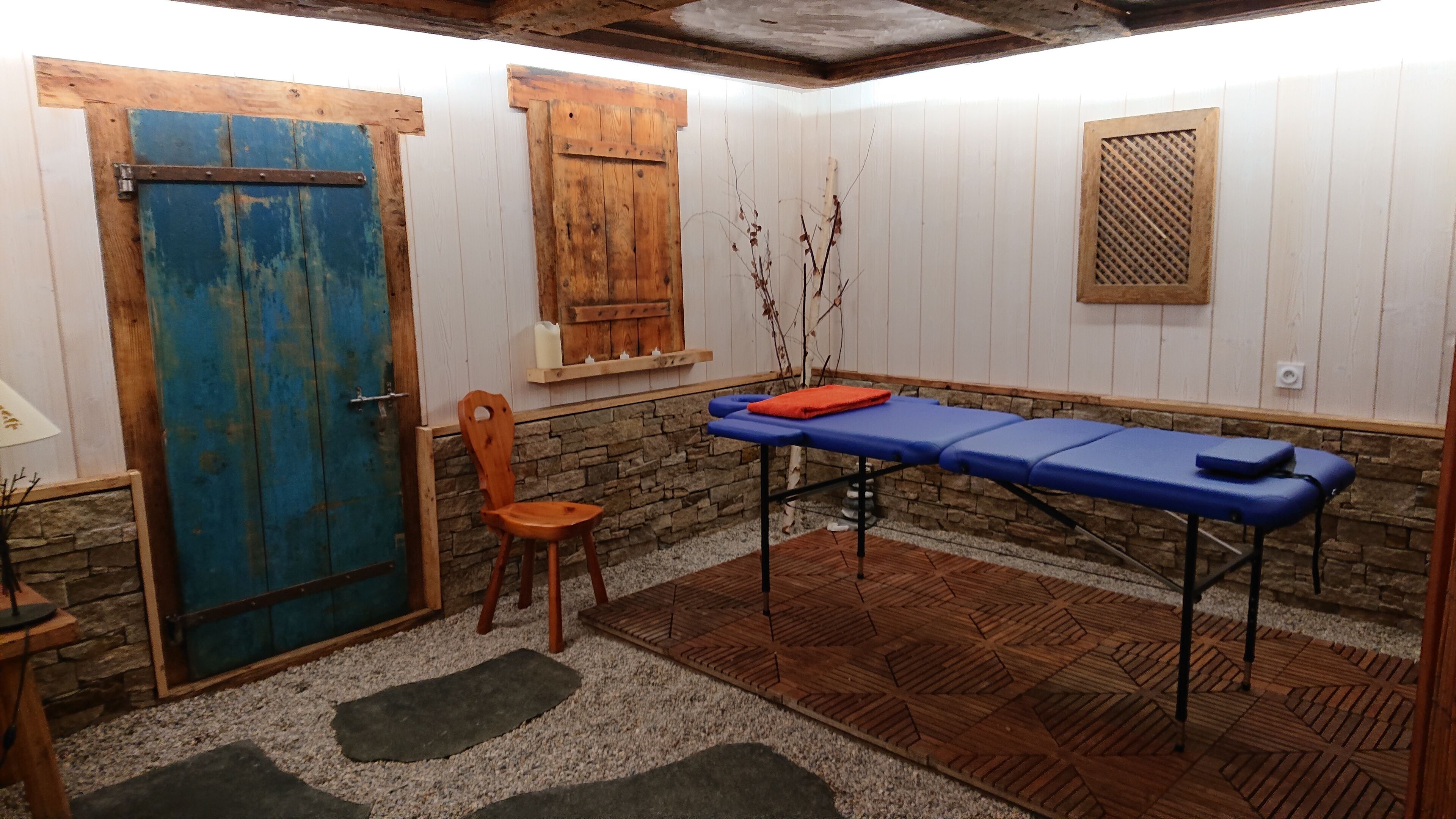 Ein Zimmer mit blauer Tür, einem Holzstuhl, einer Massageliege und einer Steinwand.