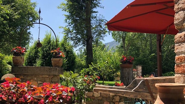 Un área de piscina exterior tranquila con un muro de piedra, plantas en macetas y un paraguas rojo.