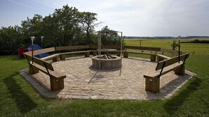 En rund ildsted i sten med bænke hele vejen rundt, beliggende i en græsbevokset park med træer i baggrunden.