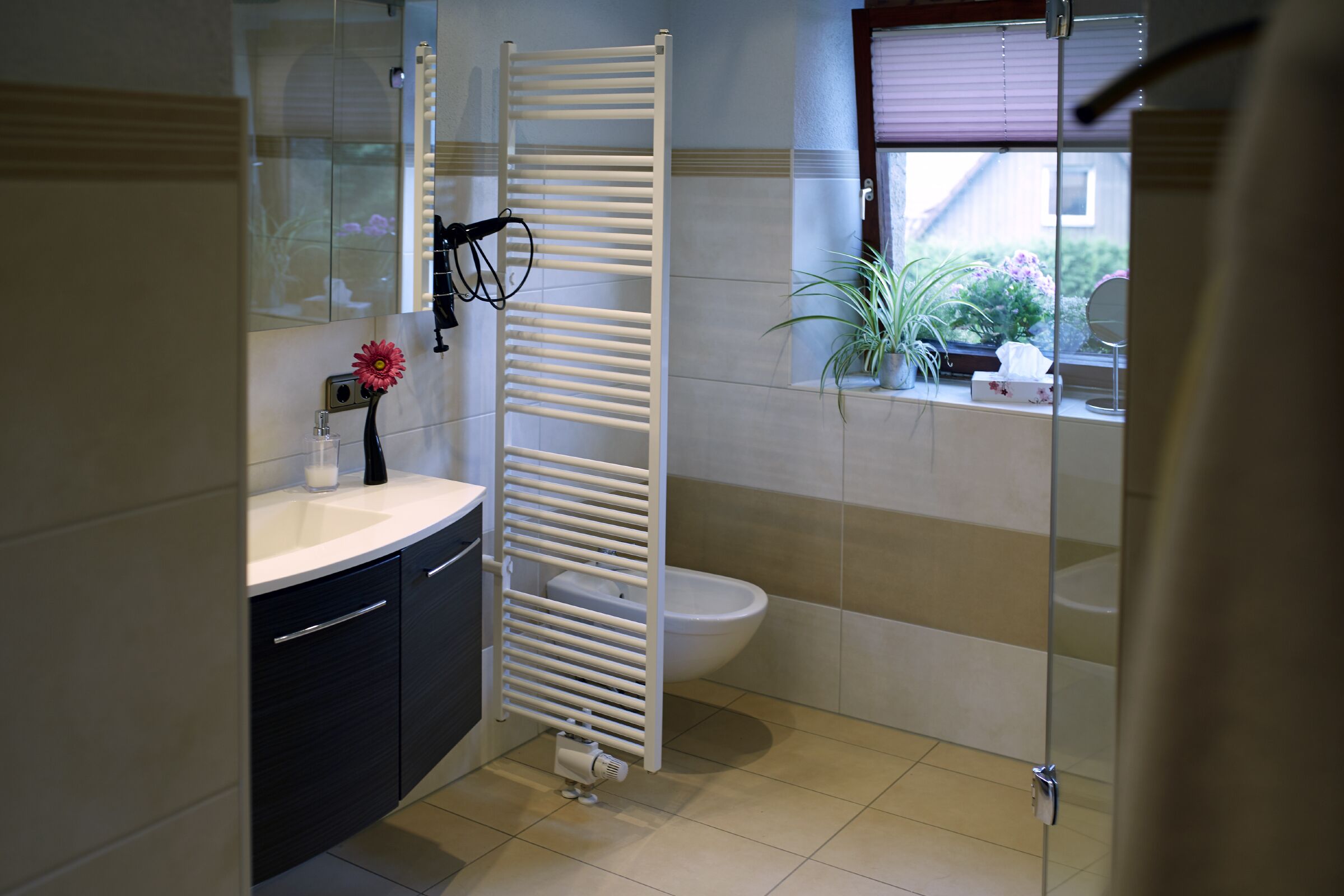 Une salle de bain moderne avec une baignoire blanche, des toilettes et un lavabo. On y trouve un porte-serviettes, un miroir et une plante en pot sur le rebord de la fenêtre.