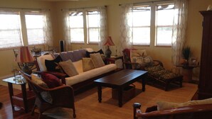 Una sala de estar con un sofá, sillones, una mesa de centro y una mesa de comedor. Hay ventanas con cortinas, un ventilador de techo y una lámpara sobre una mesita auxiliar.