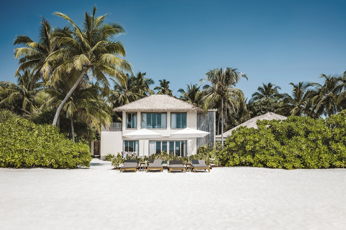 A beachfront property with a thatched-roof building, lounge chairs, and palm trees.