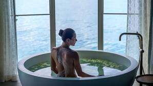 A person in a bathtub with a view of the sea through large windows.