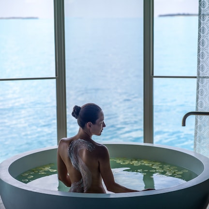 A person in a bathtub with a view of the sea through large windows.