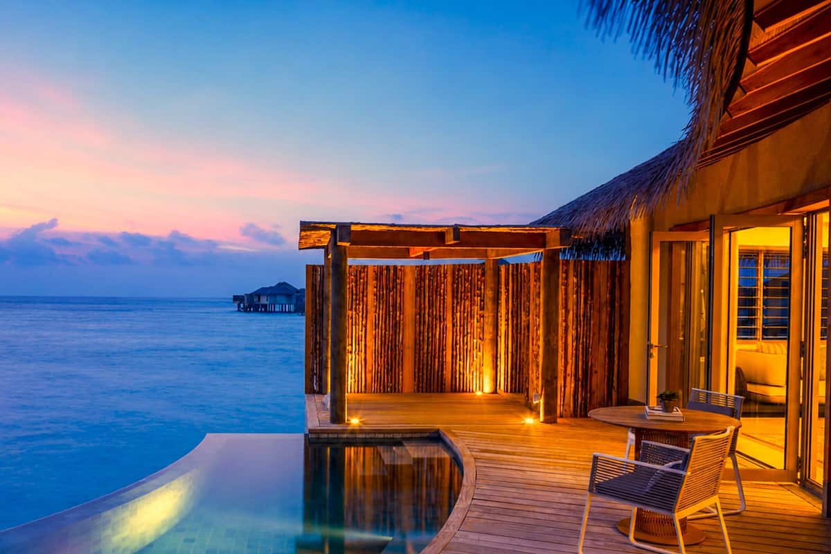 A infinity pool with a wooden deck, a dining area with chairs, and a view of the ocean and overwater bungalows.
