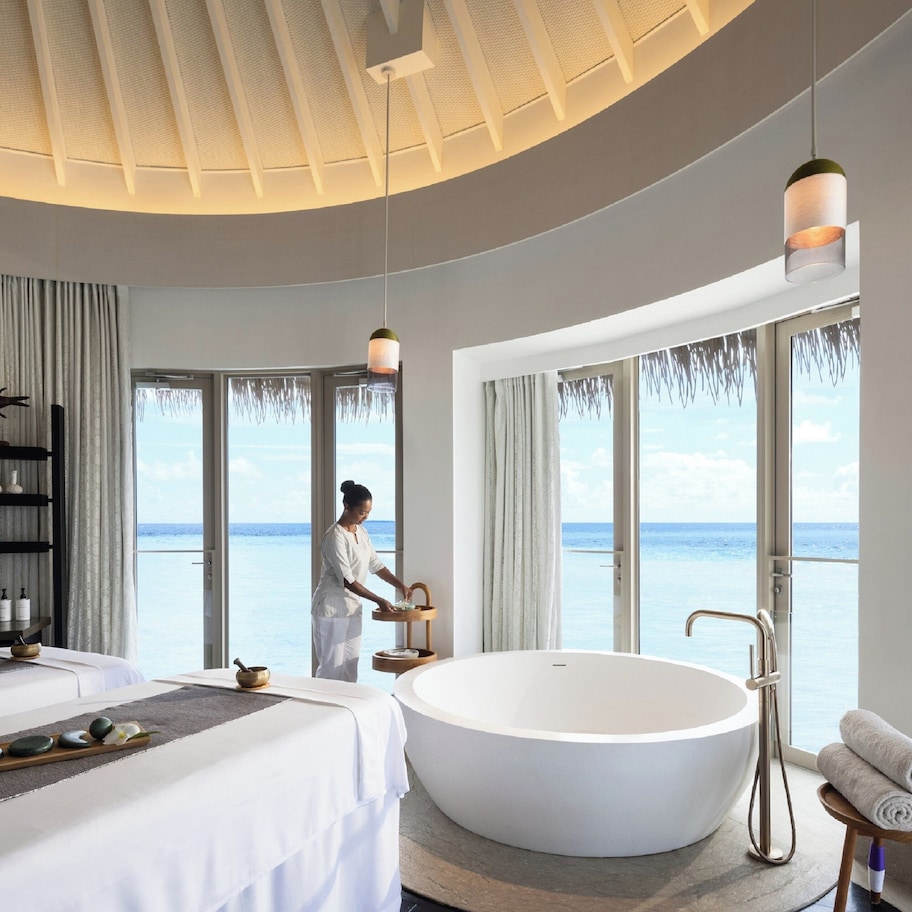 A modern hotel room with a large bathtub, a bed, a vanity with toiletries, and a view of the sea.