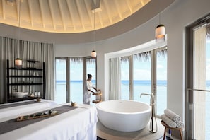 A modern hotel room with a large bathtub, a bed, a vanity with toiletries, and a view of the sea.