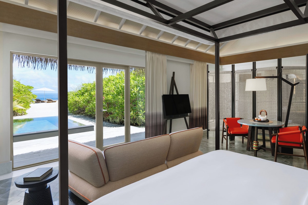 A modern hotel room with a large bed, a dining area with red chairs, a view of the ocean, and a balcony.