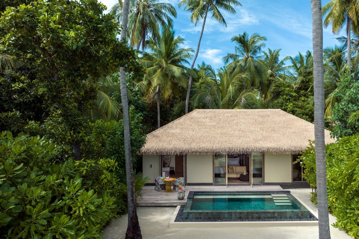 A thatched-roof bungalow with a pool, surrounded by lush greenery and palm trees.