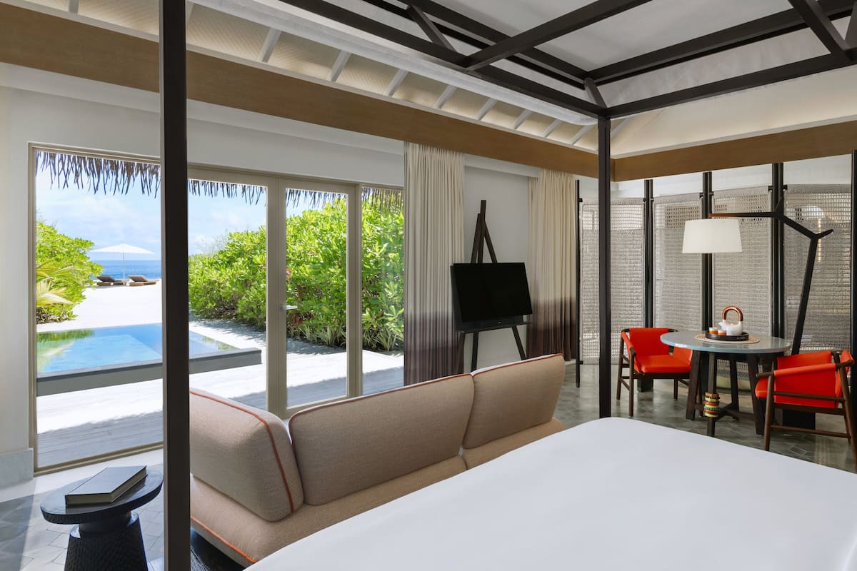 A modern hotel room with a large bed, a sofa, a dining table with chairs, a painting on easel, and a view of the ocean.