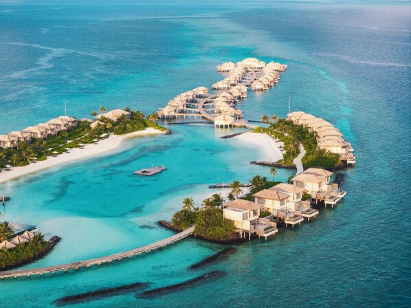 An aerial view of a resort with overwater bungalows connected by walkways, a sandy beach, and clear turquoise waters.