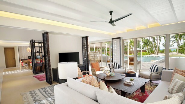 A spacious living room with a large sofa, a coffee table, and a TV. There is a balcony with a pool and palm trees visible through the large windows.