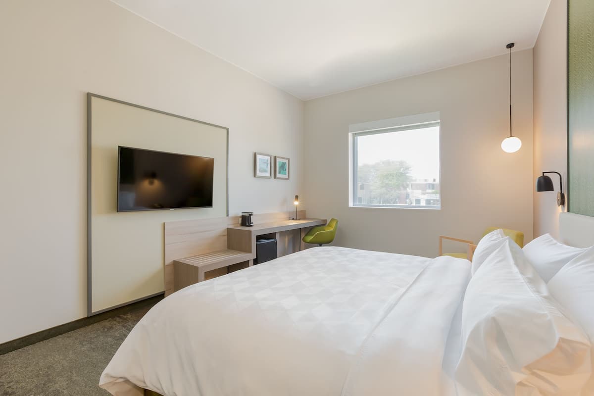 A modern hotel room with a large bed, a desk with a chair, a flat-screen TV, a window, and a small table with a lamp.