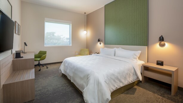 A modern hotel room with a large bed, a desk with a flat-screen TV, a green chair, and a window with a view of the city. - Holiday Inn Piura by IHG (Piura)