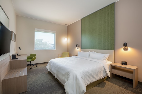 A modern hotel room with a large bed, a desk with a flat-screen TV, a green chair, and a window with a view of the city.